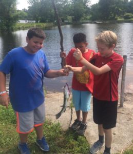 Three boys holding a caught fish by a lakeside.