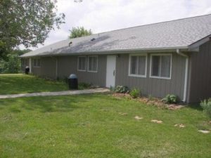 Single-story gray house with a green lawn and overcast sky.