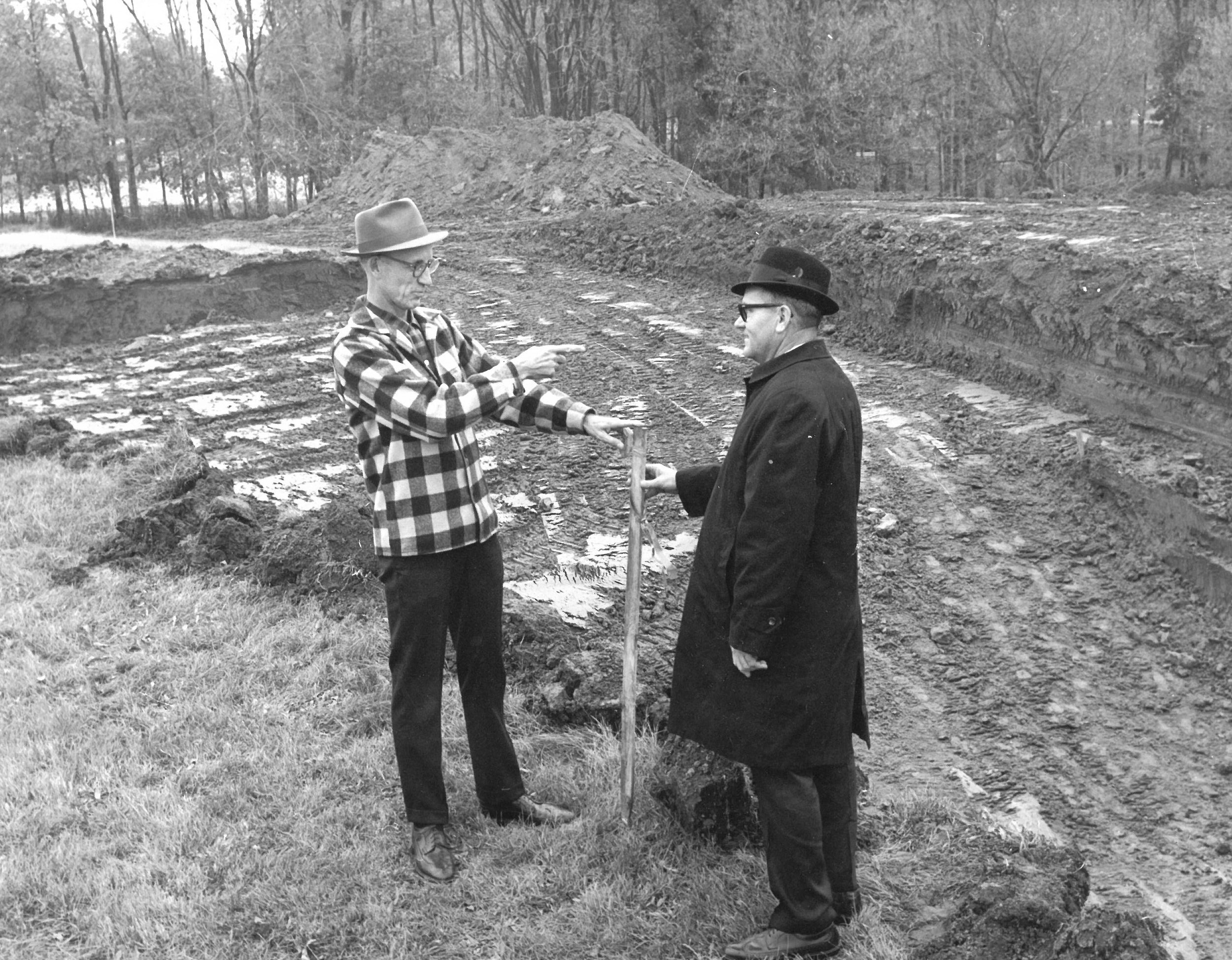 Two men discuss a construction site with one pointing and the other holding a stick.
