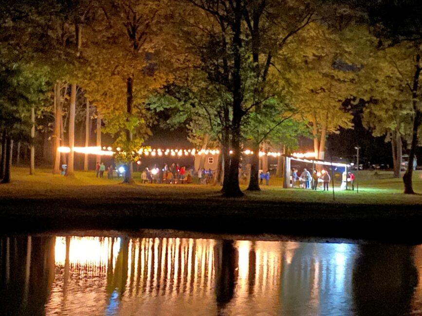 Nighttime outdoor gathering with string lights reflecting on a pond.