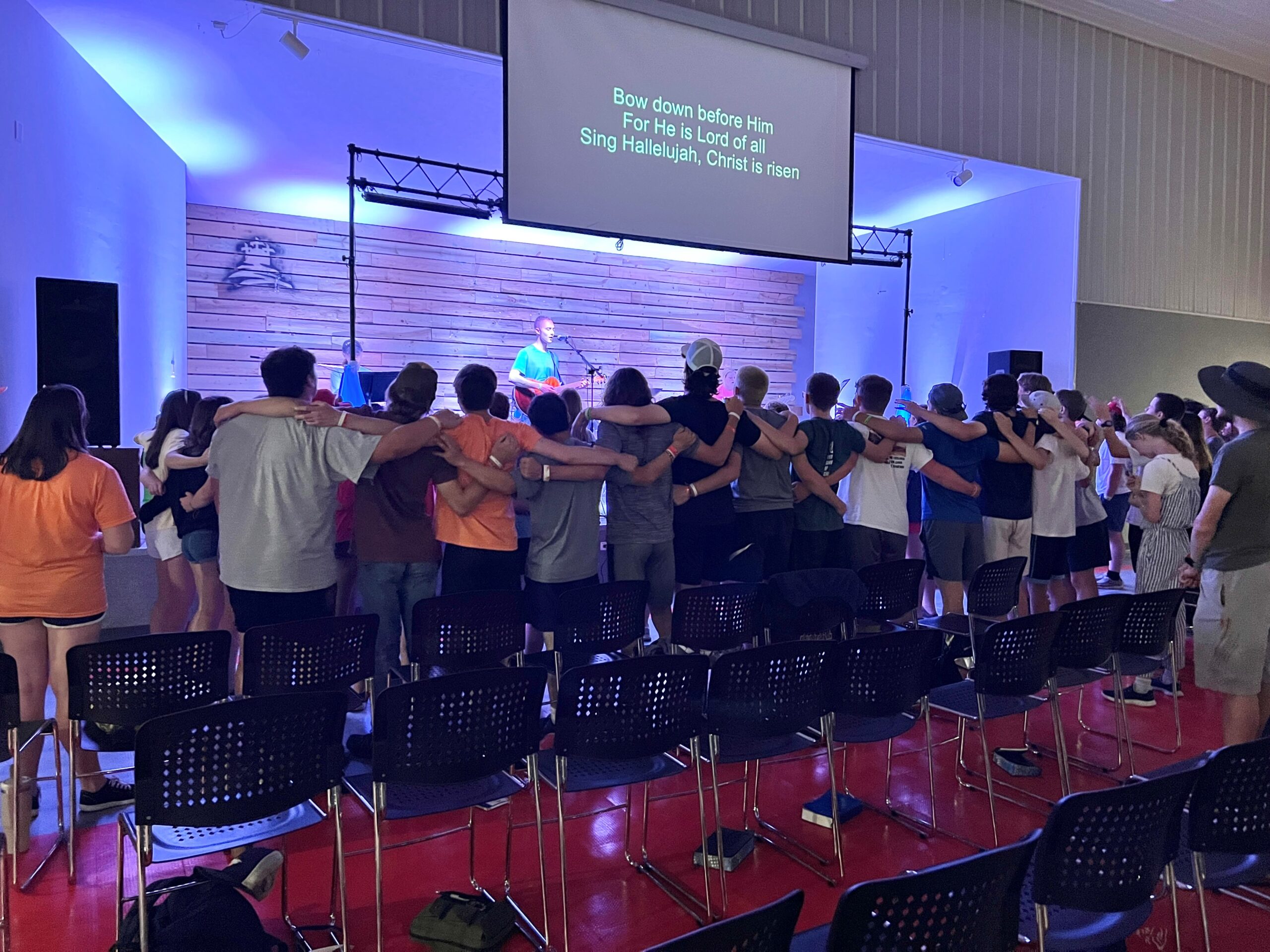 Group of people standing arm-in-arm during a worship service with song lyrics projected.