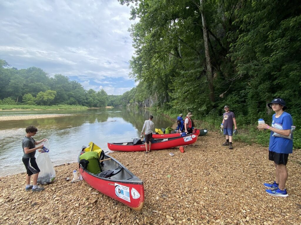 People preparing canoes near a riverbank surrounded by trees.